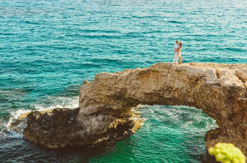 Wide shot of bridge and groom embracing on rock arch over sea