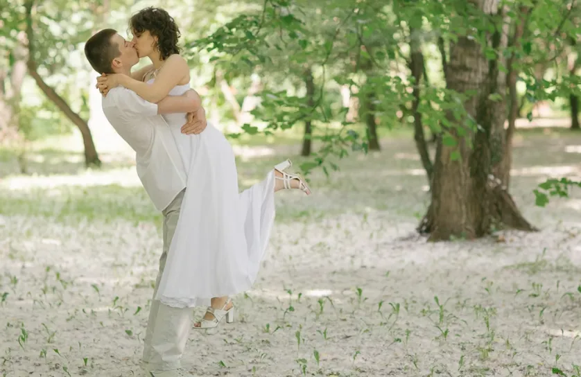 Eedding couple embracing and kissing in a natural woodland setting, captured in soft daylight