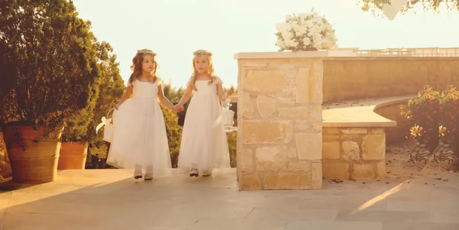 Two young children wearing white dresses holding hands and walking