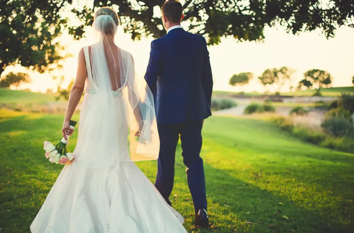 Bride and groom walk hand in hand along a grassy path with a river in the background