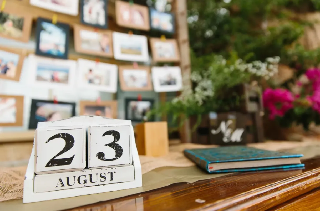 Close up of wooden wedding date calendar on the 23rd August, on a table, with guestbook and blurred guest photographs pinned to a noticeboard behind it
